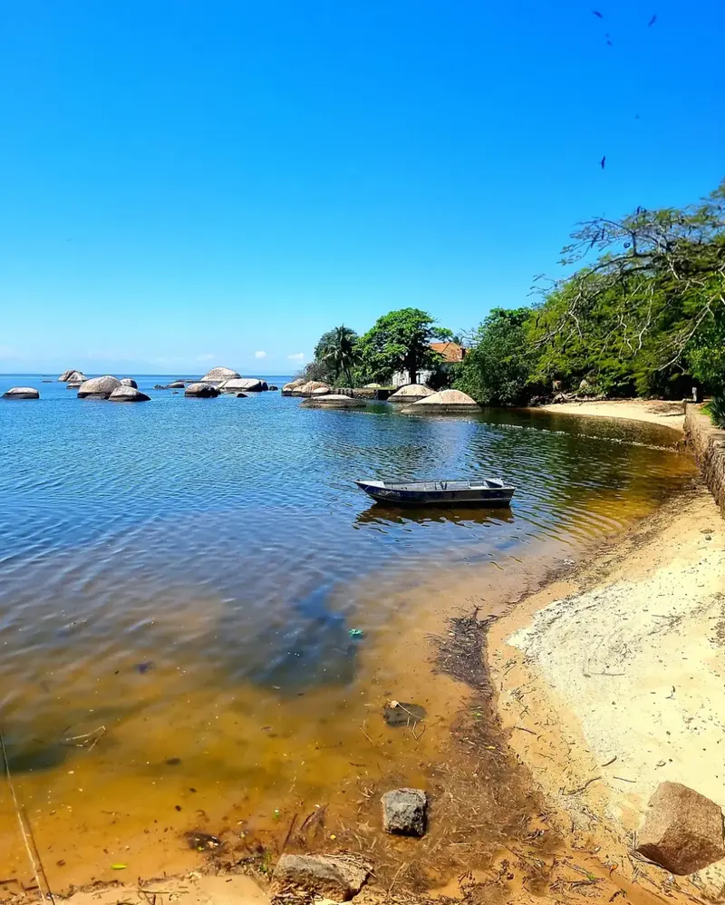 Enseada de águas calmas com pedras arredondadas, um barco pequeno e árvores à beira da praia.