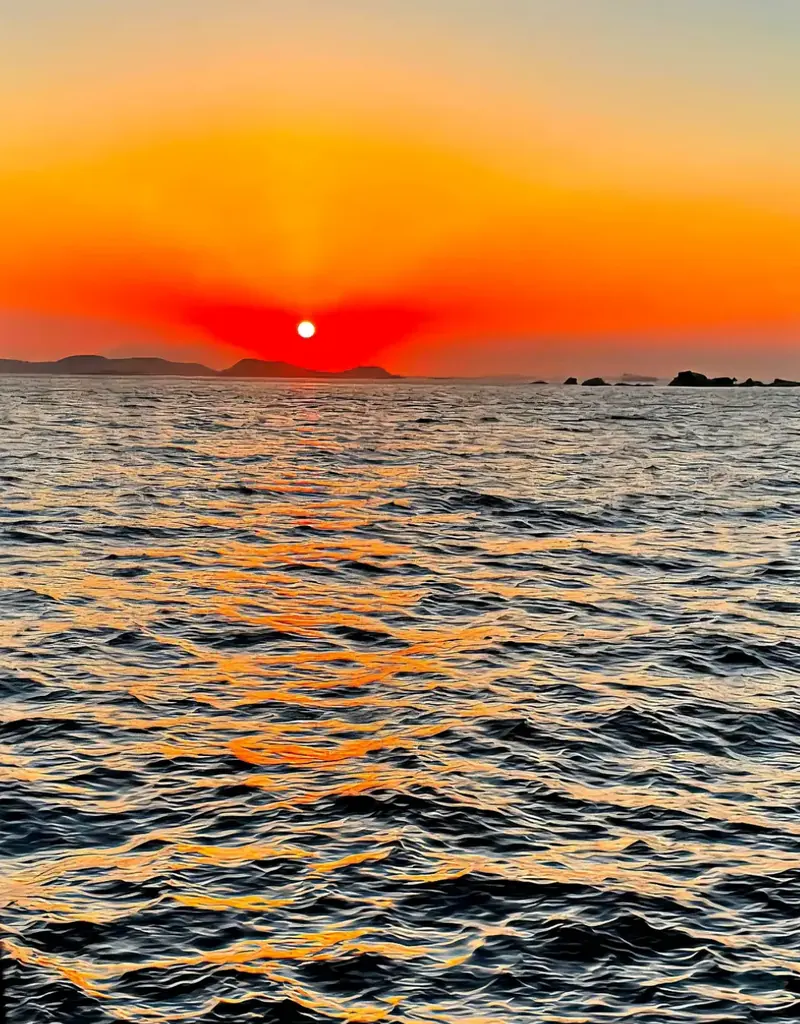 Pôr do sol alaranjado sobre o mar, com a luz refletida nas ondas e morros baixos no horizonte.