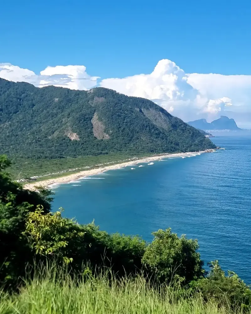 Praia de areia clara ao pé de um morro coberto de mata, vista do alto de uma trilha, com o mar azul e a Pedra da Gávea ao fundo.