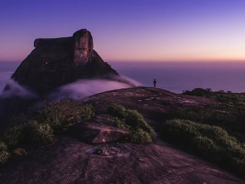 Cume arredondado de rocha ao entardecer, com a Pedra da Gávea ao fundo e nuvens baixas sobre o mar.